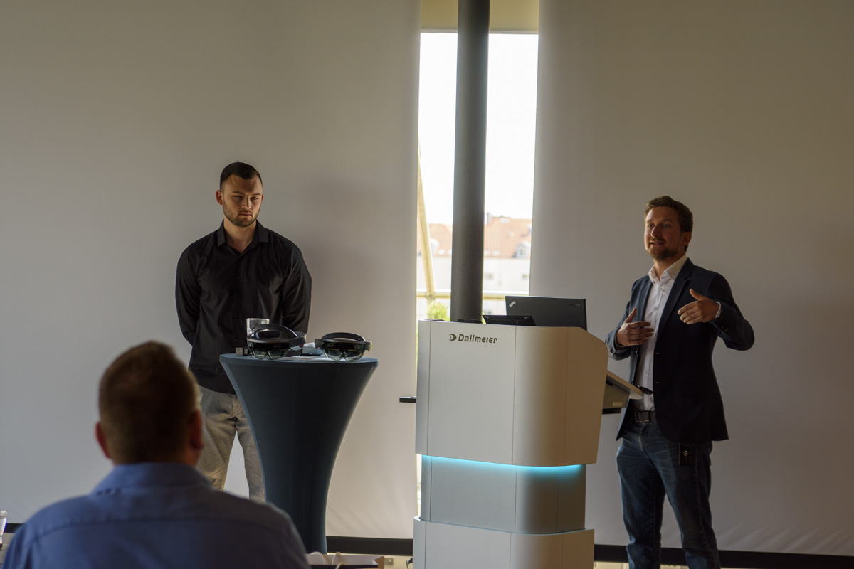 Two men are standing in front of a presentation setup. One is speaking while gesturing, and the other is listening. An audience is visible in the foreground.