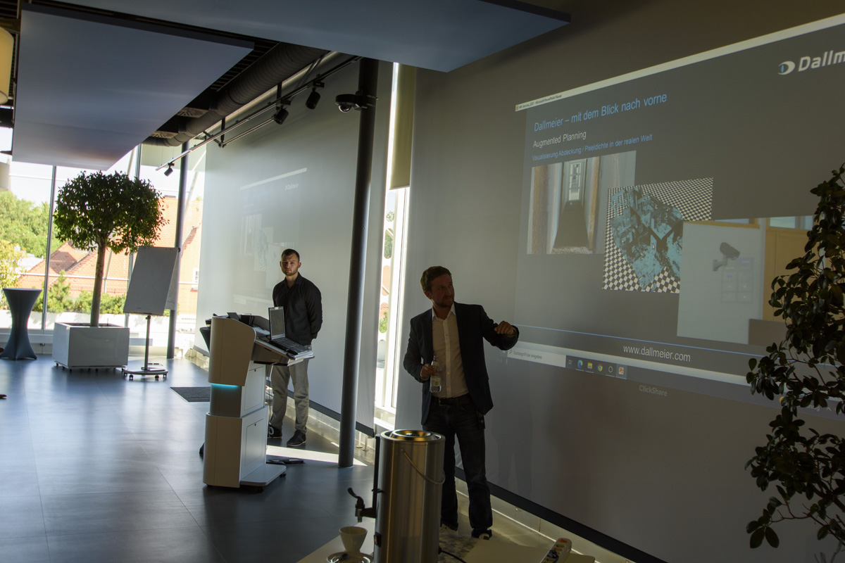 A presentation room with two speakers. One stands at a podium gesturing, while the other is next to him. A large screen displays a presentation.
