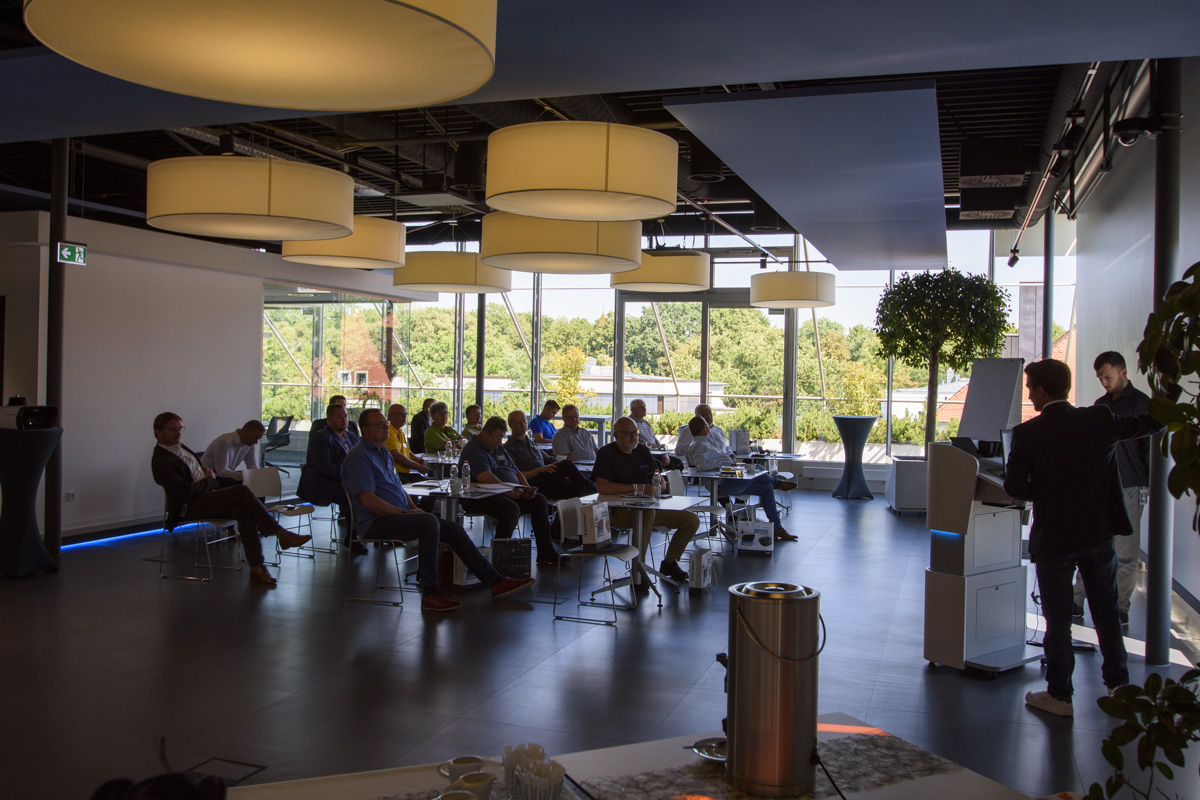 A group of people seated in a modern conference room, with large windows showcasing greenery outside. One person is presenting at the front.
