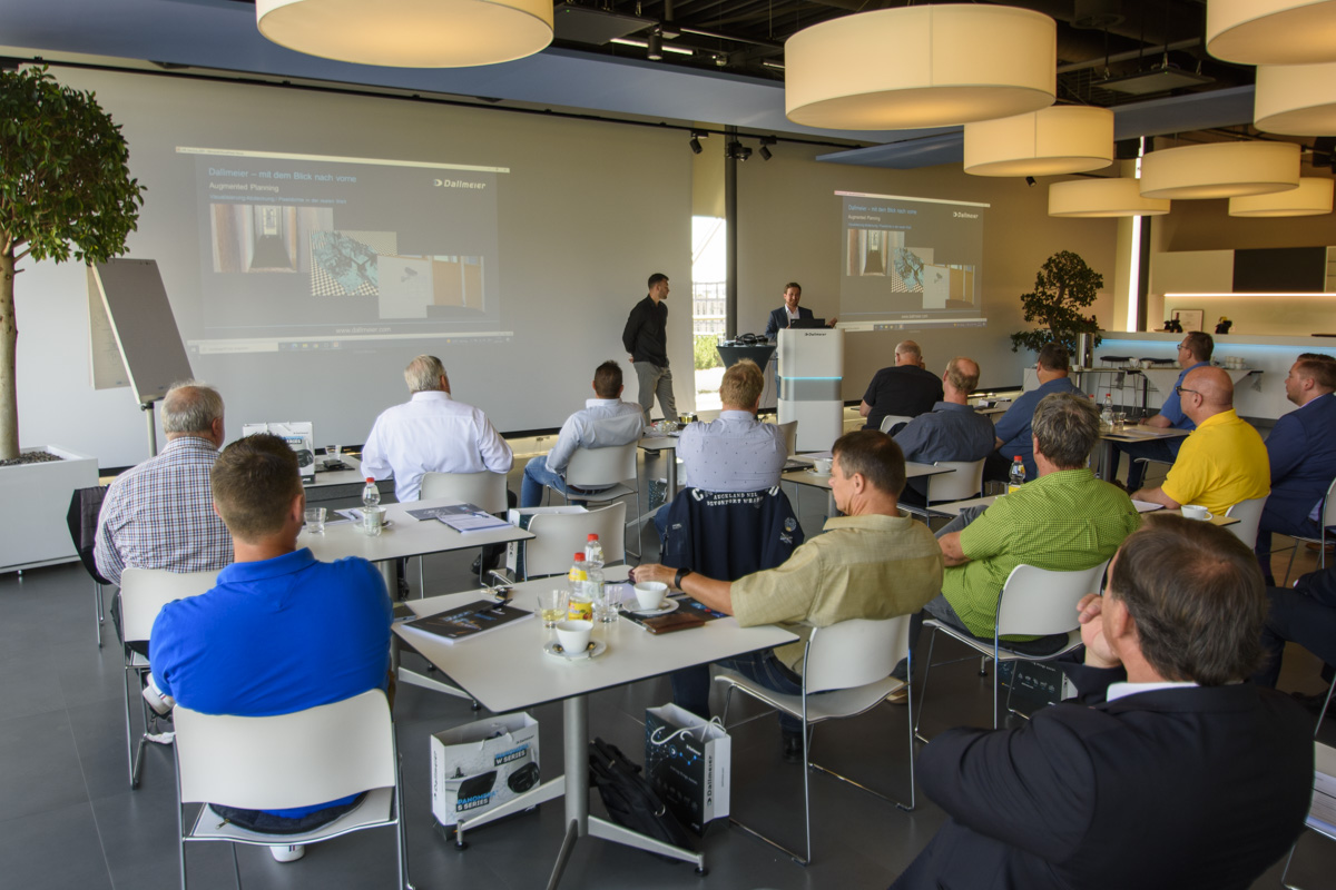 A group of people seated in a modern conference room, attentively watching a presentation on large screens, with refreshments on their tables.