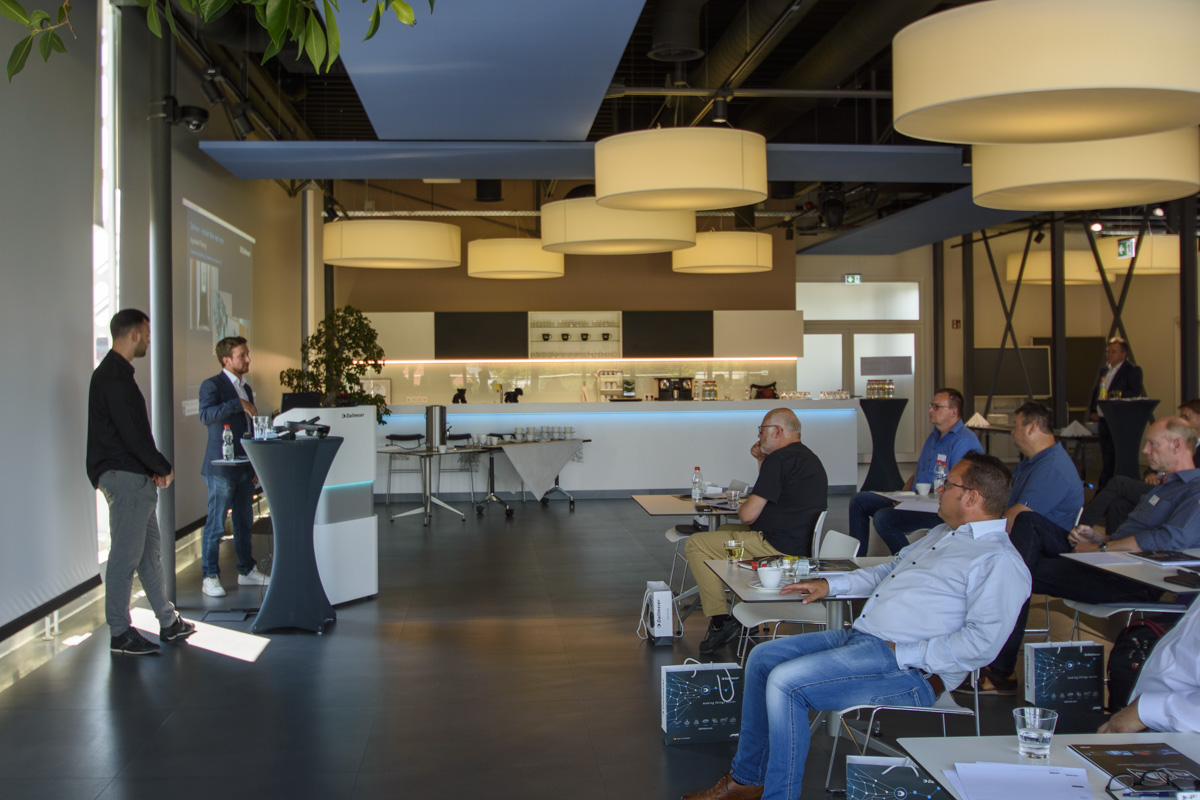 Two presenters stand at a podium addressing an audience seated in a modern conference space, with large pendant lights and a coffee area in the background.