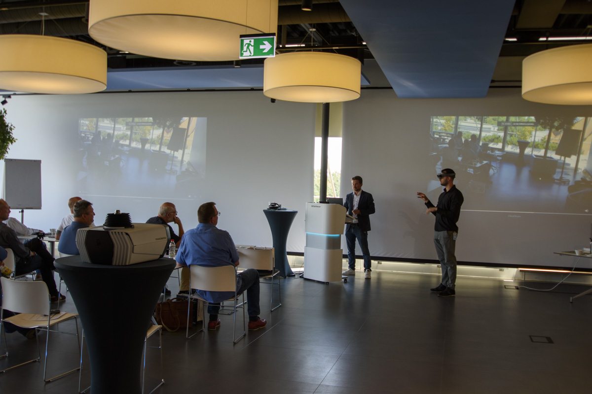 A presentation is taking place in a modern conference room with a speaker addressing an audience seated at tables. Two projectors display images on the wall.