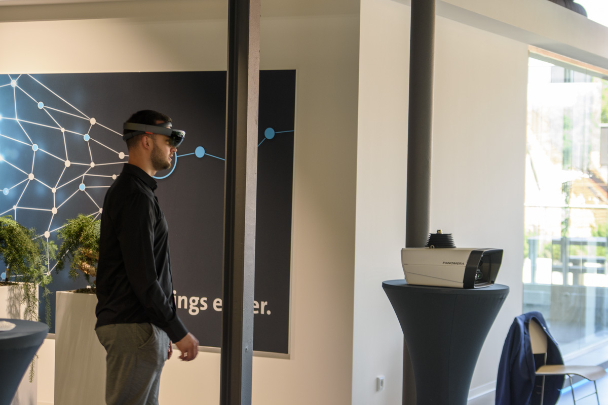 A person wearing a virtual reality headset stands in a modern room with a display featuring a network design. Nearby, a projector is placed on a table.