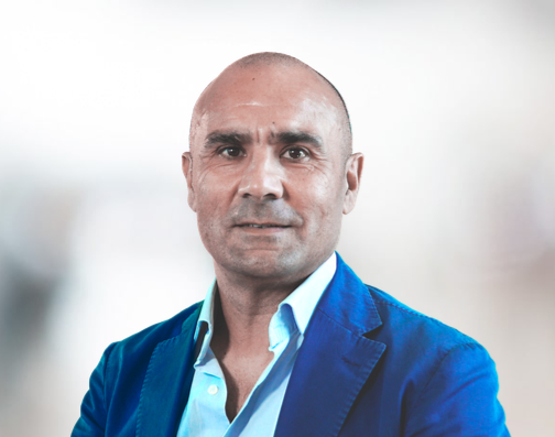 A man with short hair and a serious expression is wearing a blue blazer over a light blue shirt, standing against a soft, blurred background.