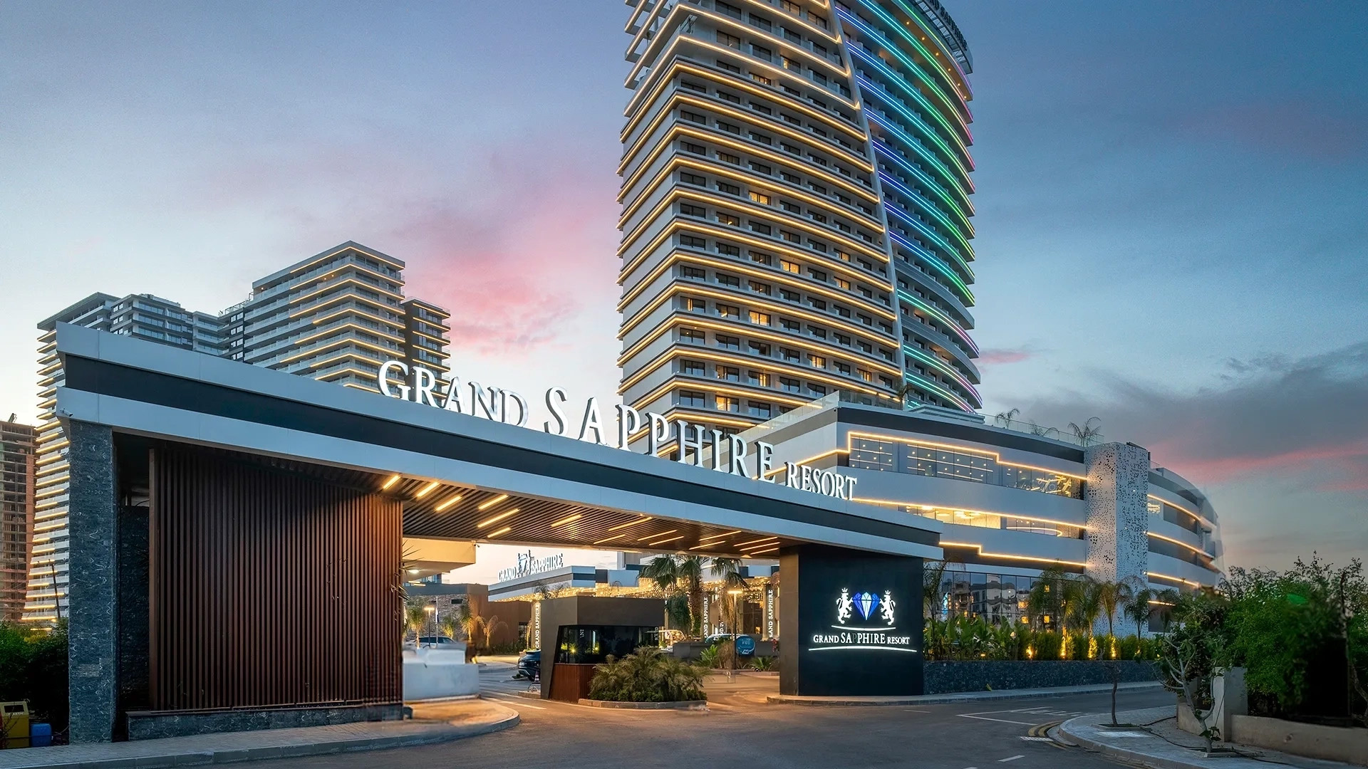 Entrance of the Grand Sapphire Resort featuring modern architecture and a welcoming sign, with colorful buildings in the background under a twilight sky.