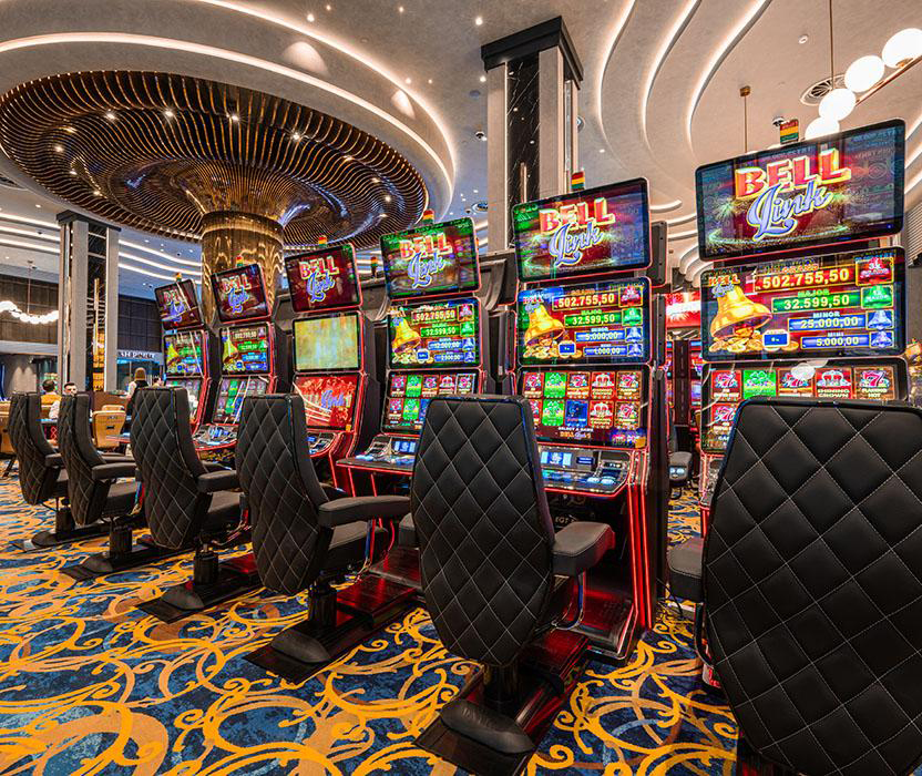 A row of colorful slot machines in a casino, featuring bright screens and plush black chairs, with decorative lighting and patterned carpets in the background.