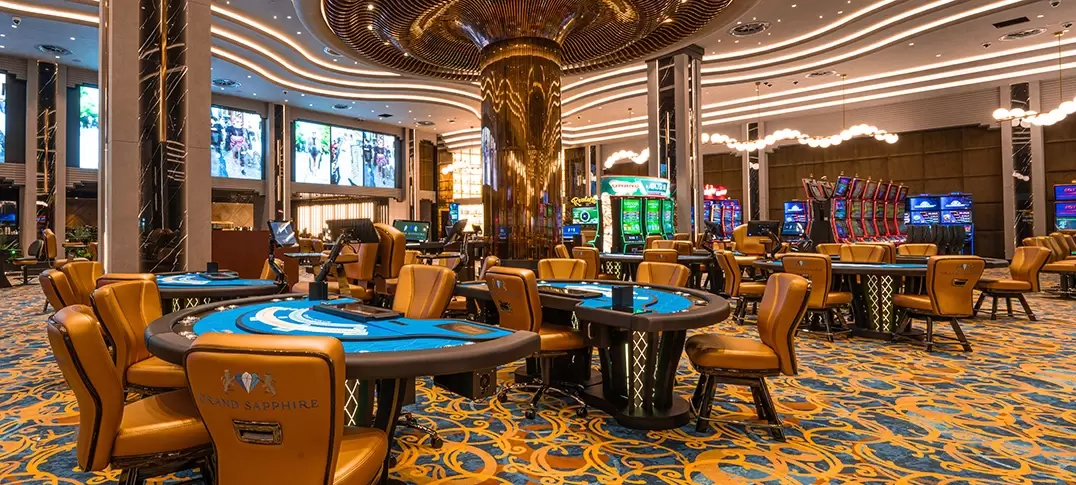 Interior of a modern casino featuring gaming tables with blue felt, golden chairs, and colorful slot machines in the background, illuminated by bright lights.