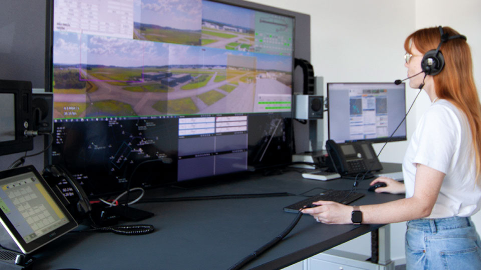 Young woman wearing a headset at her workstation with a whole view over the airport on a big screen.