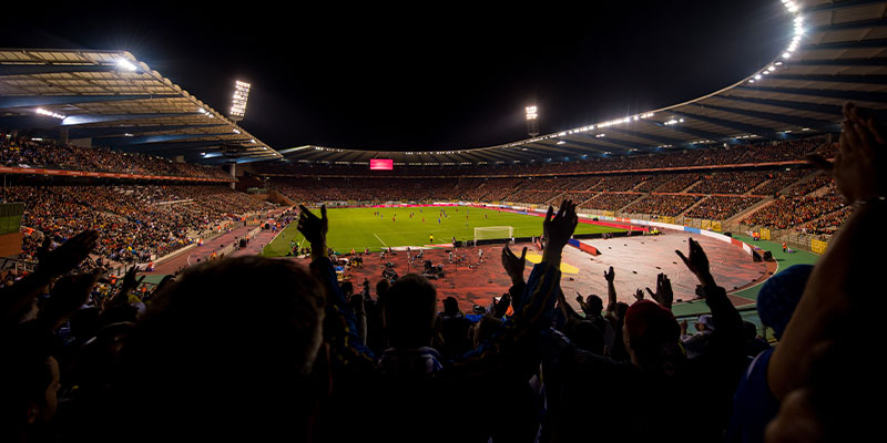 A panoramic view of a large stadium filled with cheering fans at night, with a football match taking place on the field.