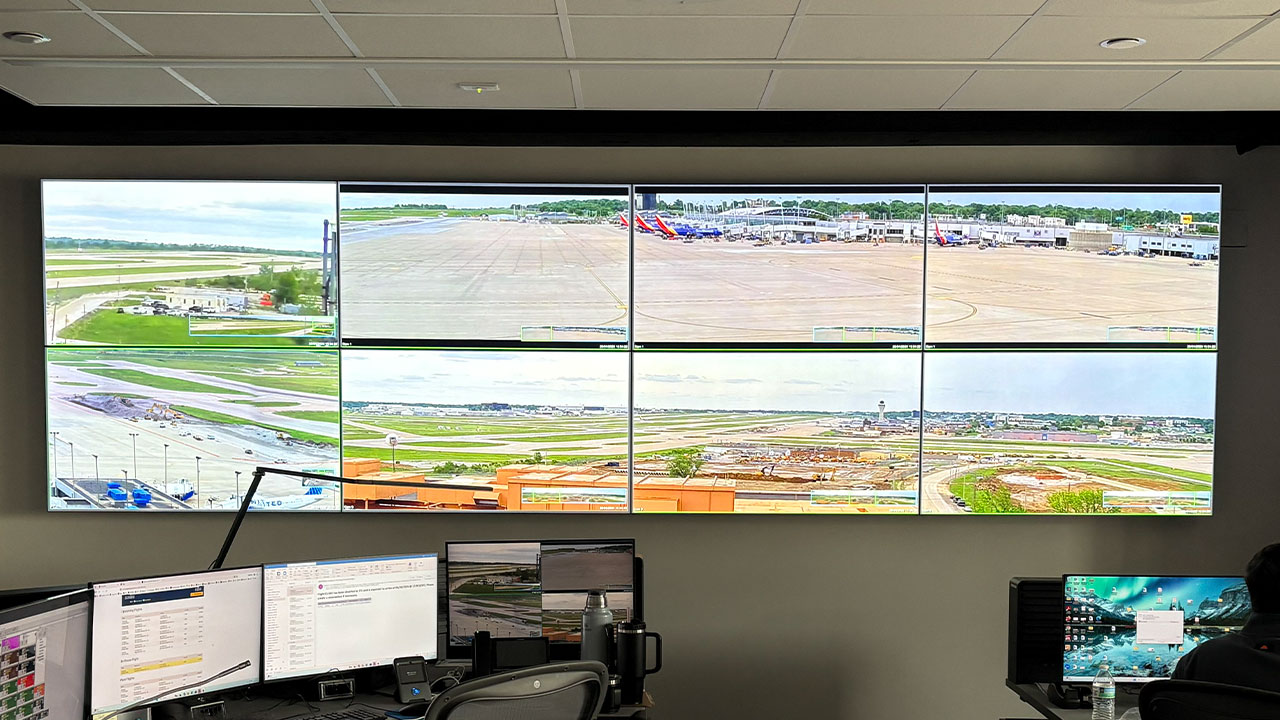 A monitoring room with large screens displaying airport views. Two operators are seated at desks, focused on their tasks, with various computer monitors showing data.