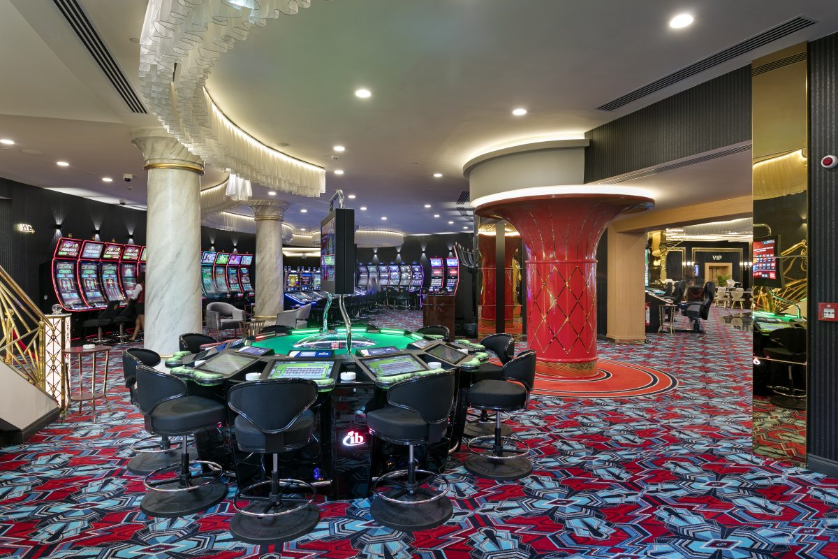 Interior of a casino featuring a gaming table surrounded by chairs, colorful carpet, and rows of slot machines in the background. Brightly lit atmosphere.