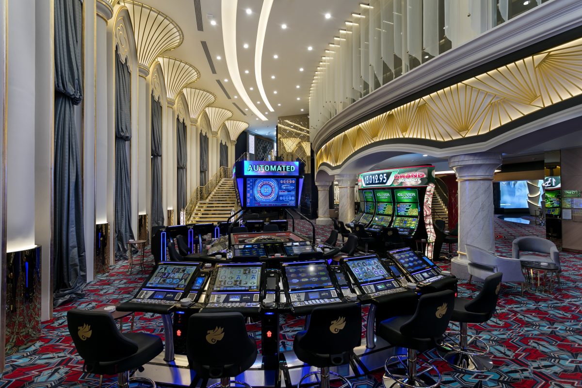 Interior of a casino featuring illuminated slot machines, gaming tables, and ornate architecture with a decorative ceiling and carpet.