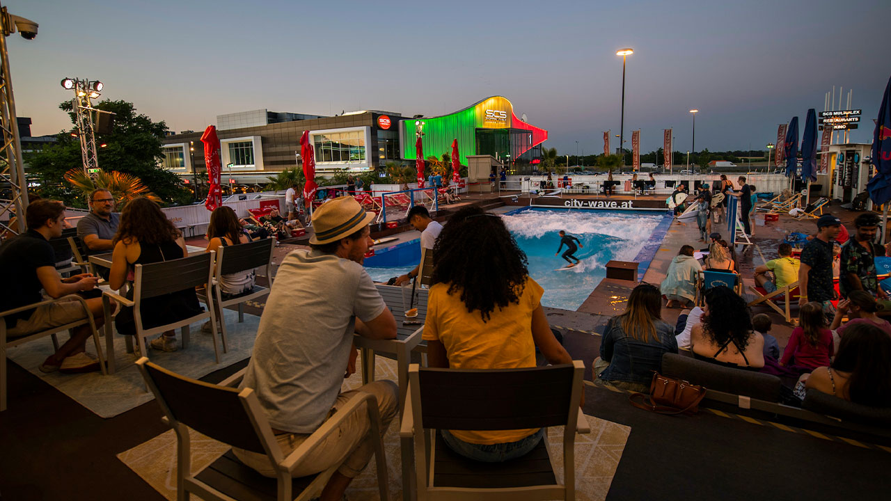 A man and a woman chatting at a table with drinks while watching surfer on an artificial surf wave.