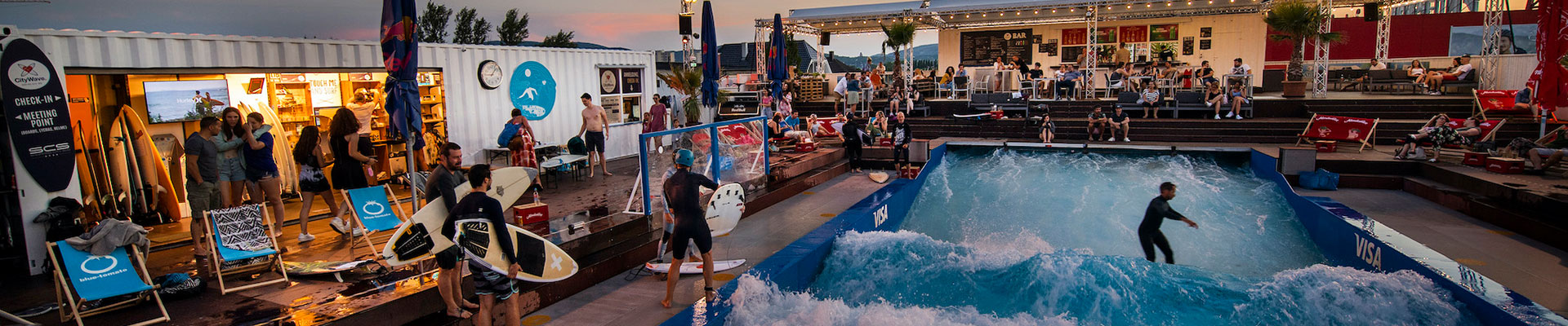 Surf wave in swimming pool with surfers and many spectators on wooden grandstands.