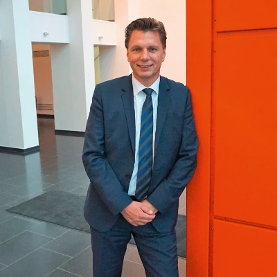 Thorsten Kinhöfer in a blue suit leans against an orange wall.